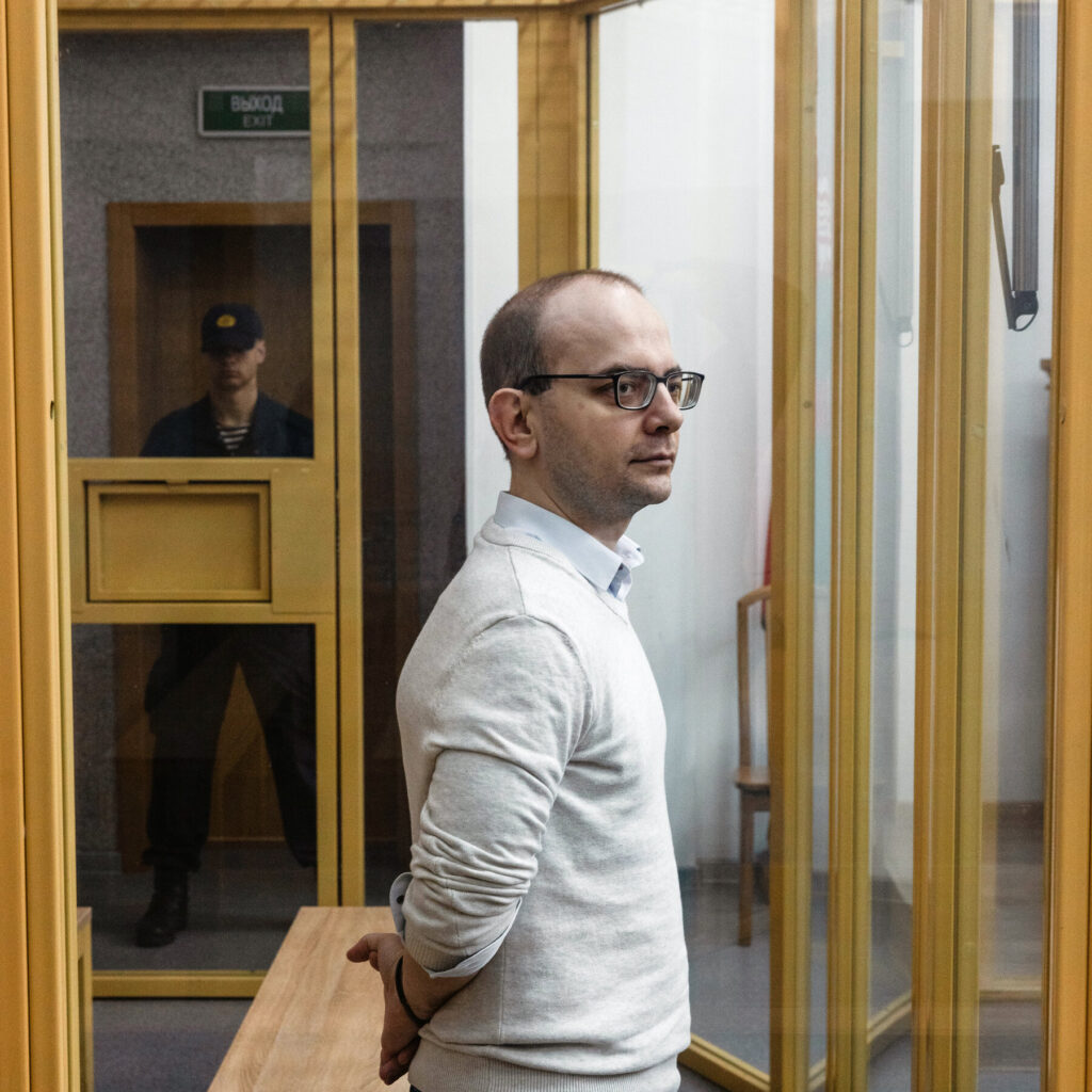 A detained individual in a glass-enclosed room, with a guard visible in the reflection, highlighting the tense situation surrounding political prisoners in Belarus.