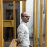 A detained individual in a glass-enclosed room, with a guard visible in the reflection, highlighting the tense situation surrounding political prisoners in Belarus.