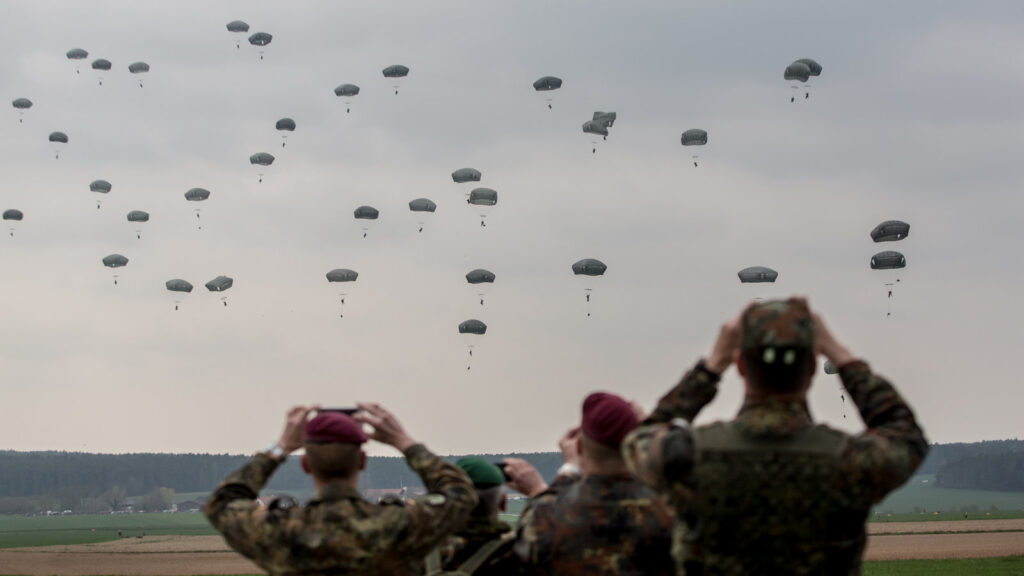 Paratroopers descend during a military exercise as observers watch from the ground, symbolizing the strategic and operational dynamics at play in NATO's Eastern flank.