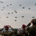 Paratroopers descend during a military exercise as observers watch from the ground, symbolizing the strategic and operational dynamics at play in NATO's Eastern flank.